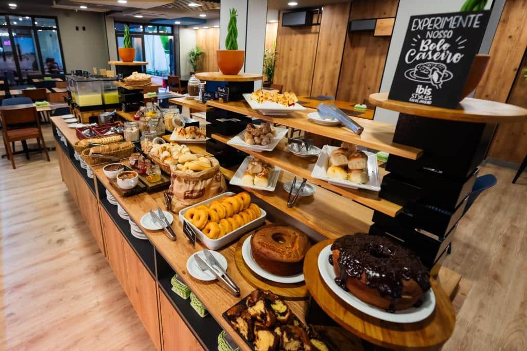 Um bufê de café-da-manhã do ibis Styles Rio de Janeiro Botafogo com doces variados, pães, bolos e frutas exibidos em prateleiras de madeira em um ambiente de restaurante.