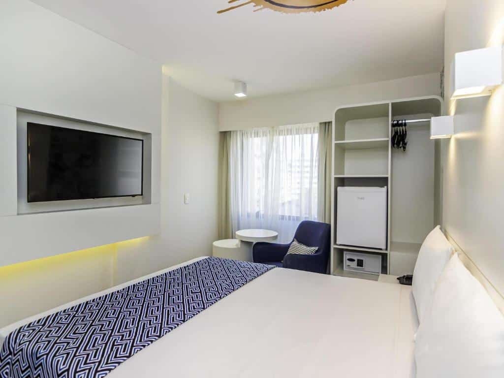 Quarto do ibis Styles Rio de Janeiro Botafogo com uma cama, TV montada, poltrona, mesa pequena, armário aberto e janela com cortinas. Essa é a uma das opções de onde ficar no Rio de Janeiro.
