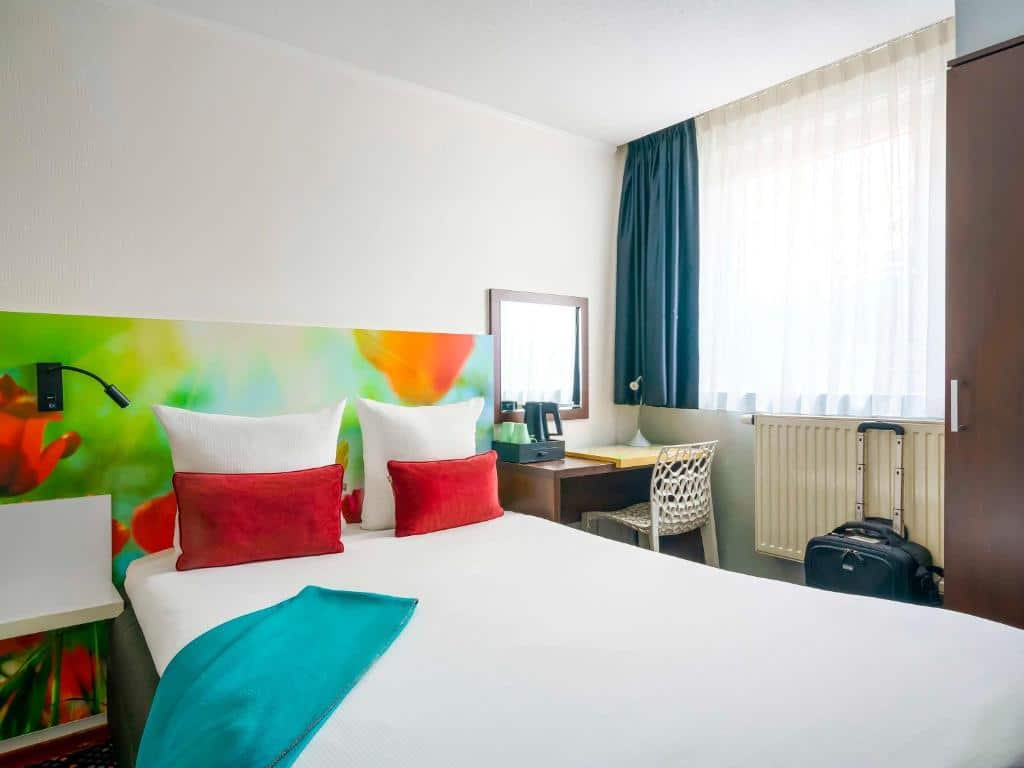 Foto do ibis Styles Amsterdam City que ilustra onde ficar em Amsterdam. Um quarto de hotel com uma cama de casal, cabeceira colorida, escrivaninha, cadeira, janela com cortinas e uma mala ao lado do guarda-roupa.