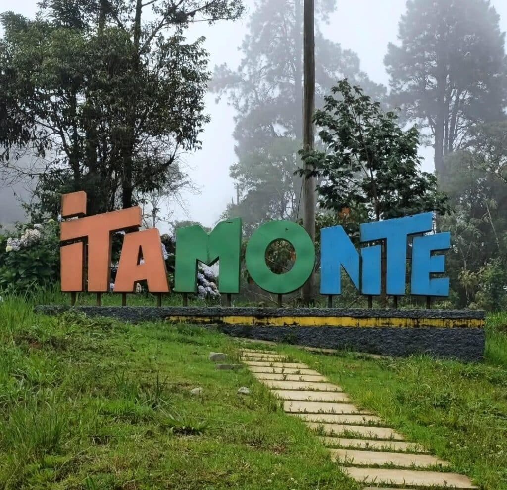 Grandes letras coloridas com a inscrição "Itamonte" em uma área gramada com árvores e neblina ao fundo.