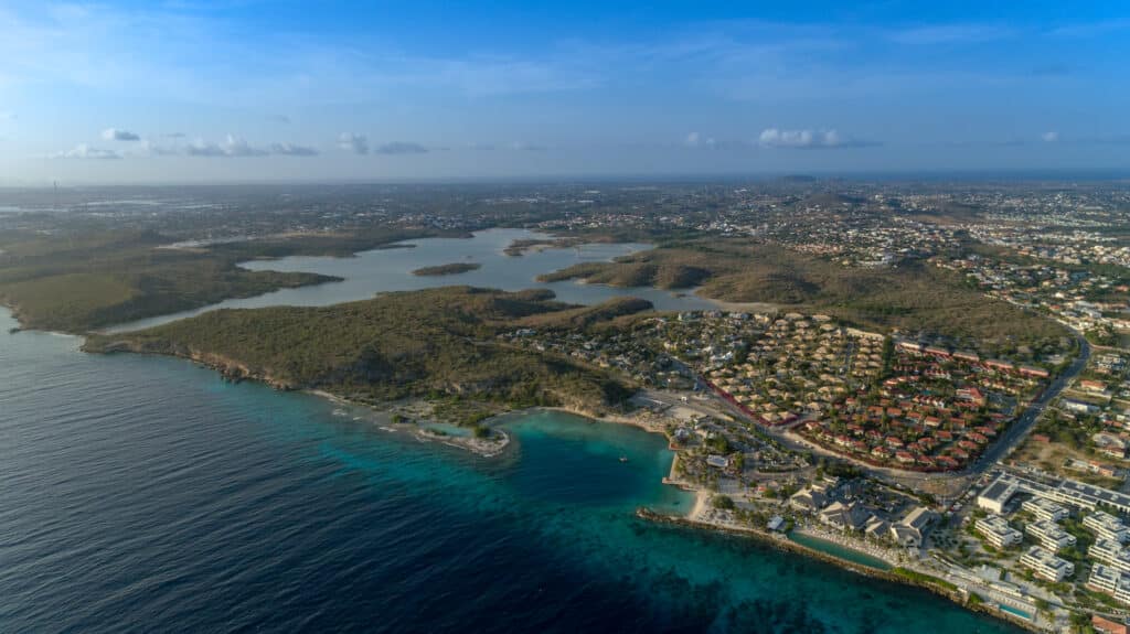 Vista aérea de Jan Thiel, em Curaçao, com água azul clara, colinas verdes e edifícios espalhados sob um céu azul.