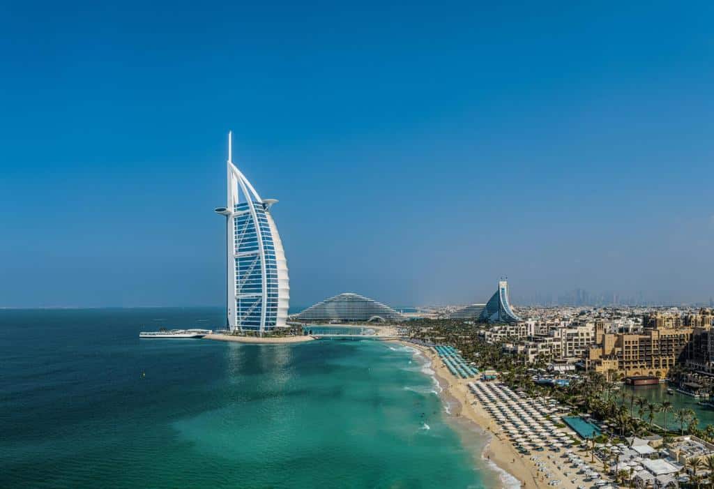 Vista aérea do hotel Burj Al Arab na costa de Dubai, com uma praia de areia e água azul clara.
