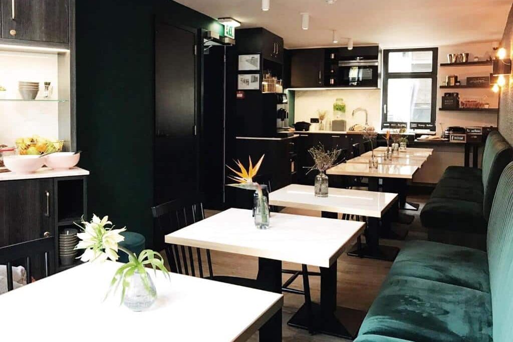 Foto do Linden Hotel. Interior de cafeteria moderna com mesas brancas, bancos verdes almofadados, cadeiras pretas e pequenos vasos de flores sobre as mesas.