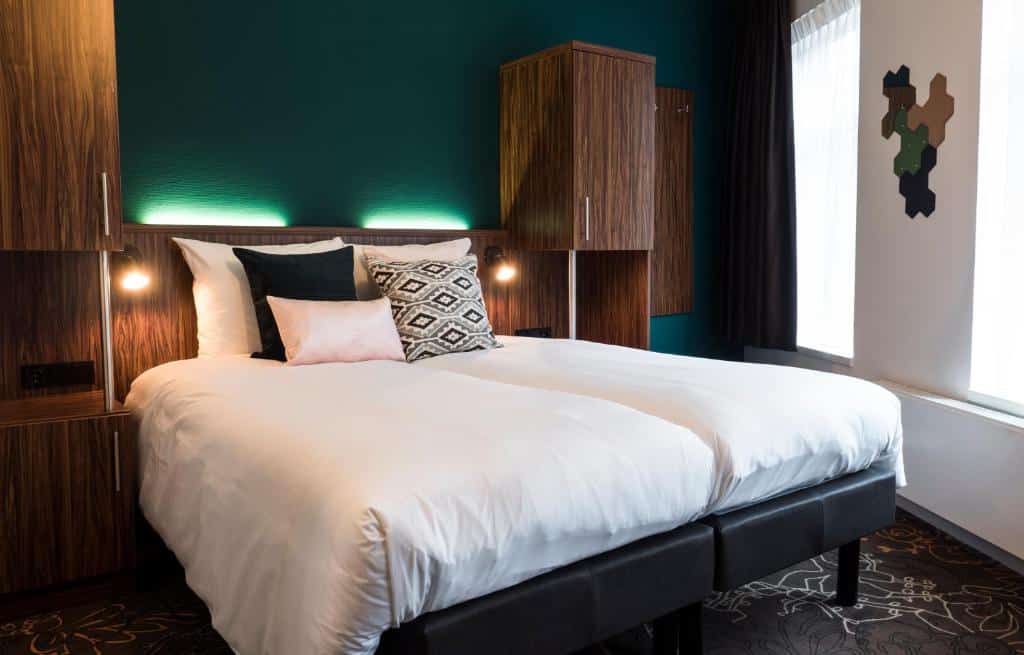 Foto do Linden Hotel que ilustra onde ficar em Amsterdam. Quarto de hotel moderno com uma cama de casal, móveis de madeira escura, travesseiros de destaque e luz natural de duas janelas.
