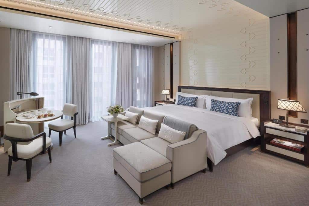 Quarto do Mandarin Oriental com uma cama grande, uma área de estar, uma pequena mesa de jantar e janelas do chão ao teto com cortinas transparentes. A foto faz parte da lista de onde ficar em Doha.