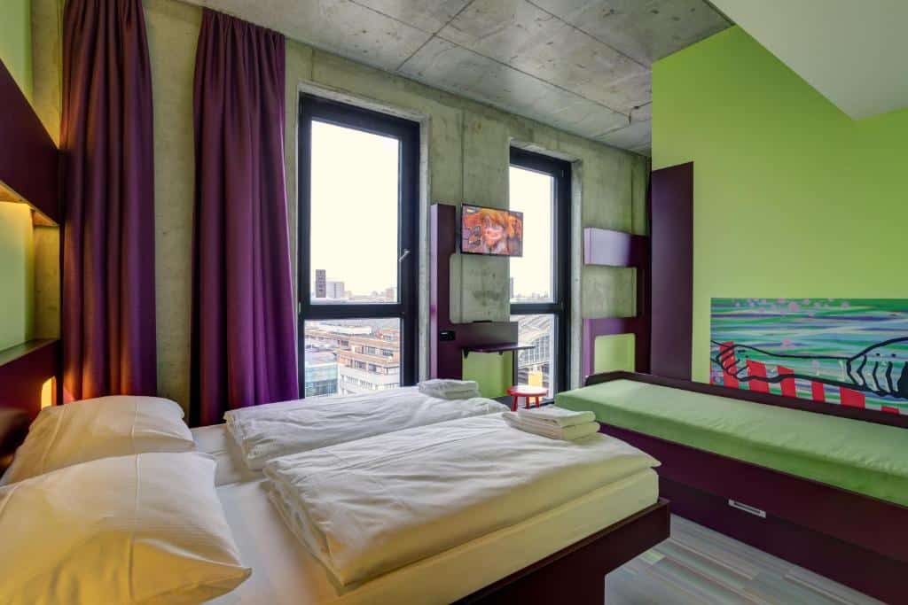 Um quarto de hotel moderno no Meininger Hotel Berlin com duas camas, janelas grandes, cortinas roxas, paredes verdes e uma TV na parede.