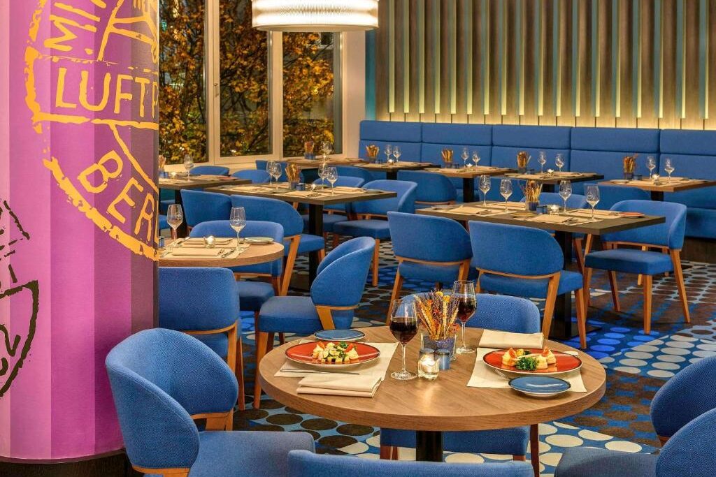Interior de restaurante moderno no Mercure Hotel Berlin com cadeiras azuis, mesas postas, piso estampado e um pilar roxo com escrita amarela.