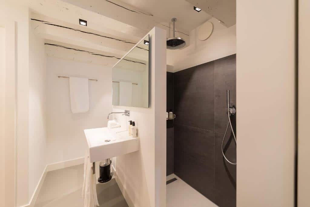 Foto do Milkhouse Luxury Stay Amsterdam. Banheiro moderno com pia branca, espelho, toalheiro e chuveiro com azulejos escuros e chuveiro com efeito de chuva.