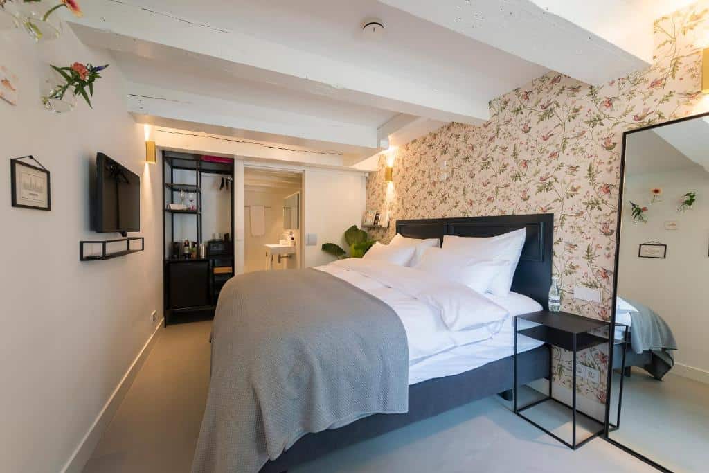 Foto do Milkhouse Luxury Stay Amsterdam que ilustra onde ficar em Amsterdam. Uma cama bem arrumada com roupas de cama brancas em um quarto moderno com papel de parede floral e uma TV na parede.