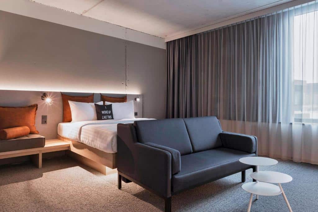 Foto do Moxy Frankfurt City Center. Quarto de hotel moderno com uma cama de casal, sofá cinza, pequenas mesas brancas e uma grande janela com cortinas transparentes.