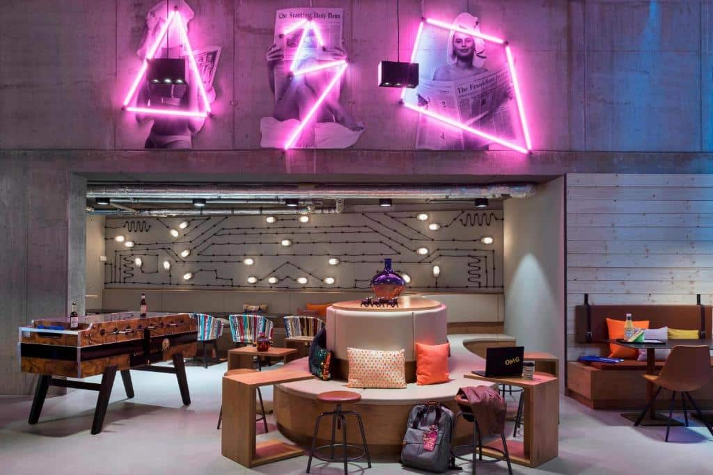 Foto do Moxy Frankfurt City Center. Lounge moderno com luzes de neon, mesa de pebolim, área de estar redonda e arte na parede em um fundo de concreto.