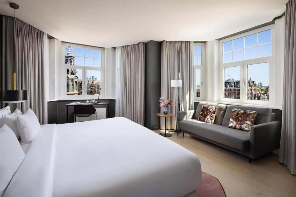 Foto do NH Collection Amsterdam que ilustra onde ficar em Amsterdam. Um quarto de hotel com uma cama grande, sofá cinza, escrivaninha e janelas que mostram uma paisagem urbana com uma torre de relógio.