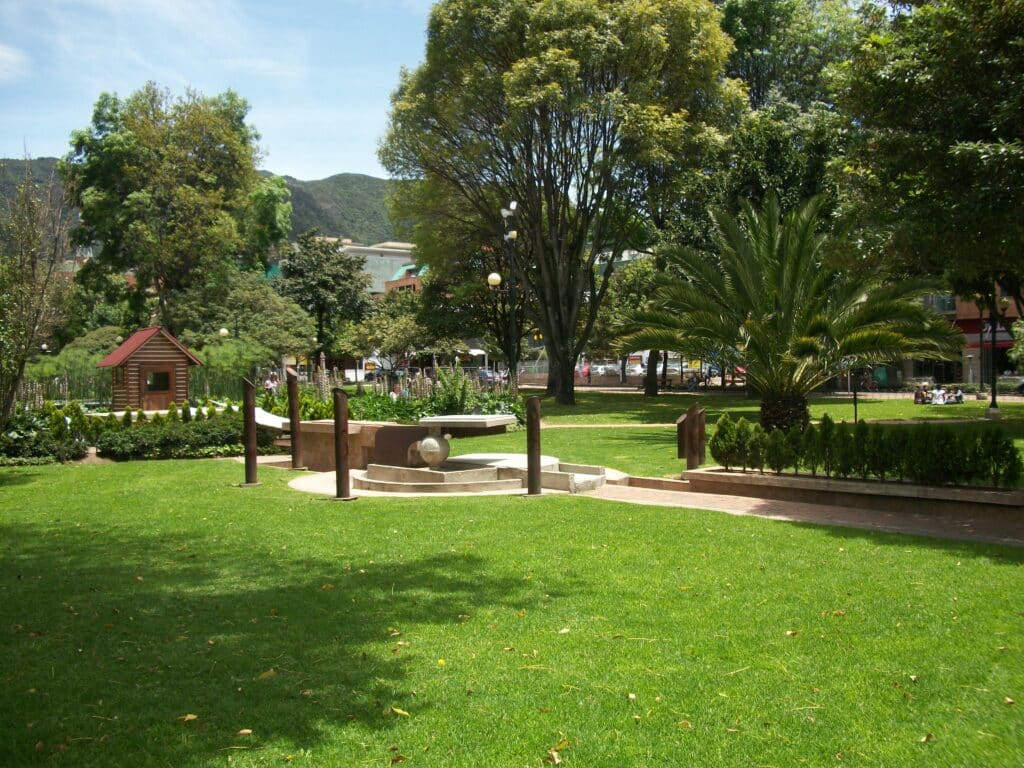 Um parque verde com árvores, grama, um pequeno playground, bancos e uma fonte circular de pedra no centro.