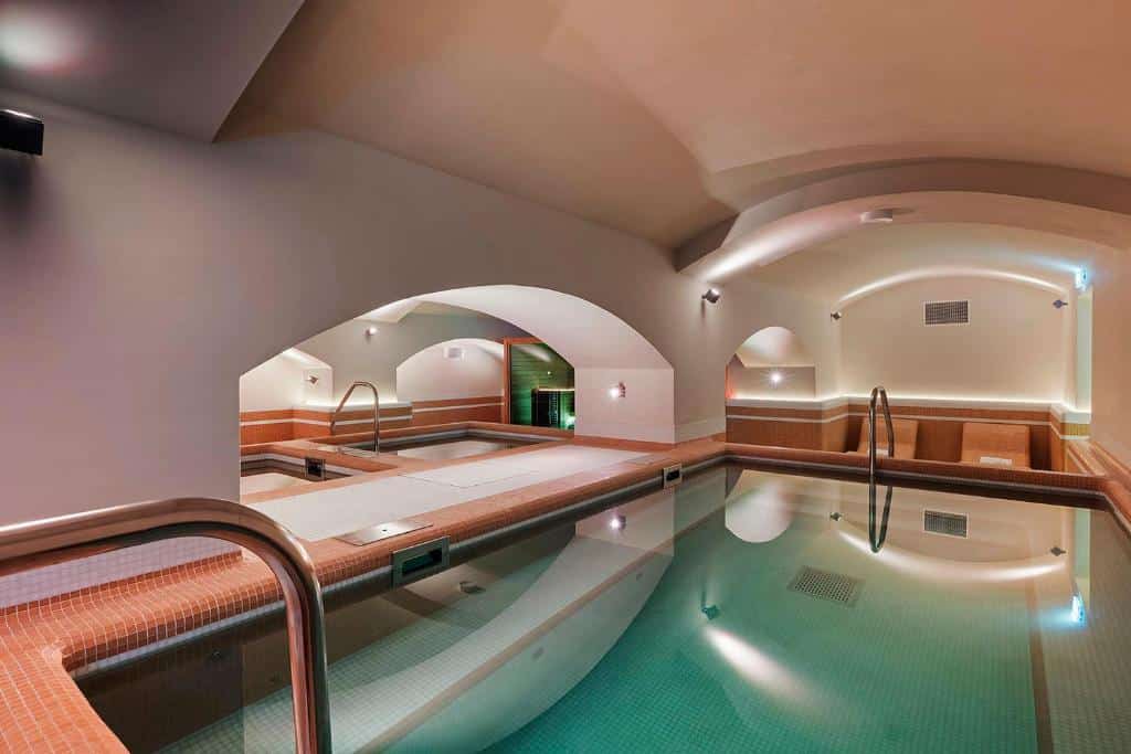 Piscina interna do Áurea Ana Palace by Eurostars Hotel Company com bordas de azulejos, tetos em arco, luzes de parede e uma pequena área de banheira de hidromassagem ao fundo.