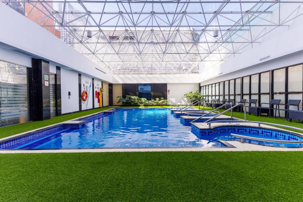 Piscina interna do Barceló Mexico Reforma com azulejos azuis, piso de grama artificial, espreguiçadeiras e um teto de vidro que permite a entrada de luz natural.