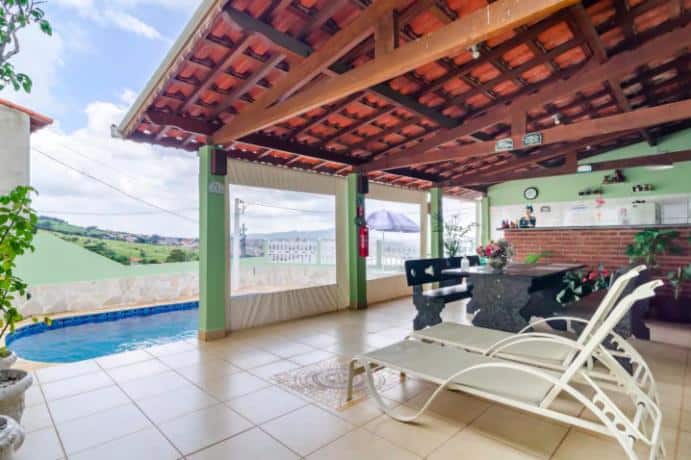 Pátio coberto da Pousada Casa da Cidinha com uma espreguiçadeira, mesa de pedra e uma pequena piscina; área da cozinha visível ao fundo.
