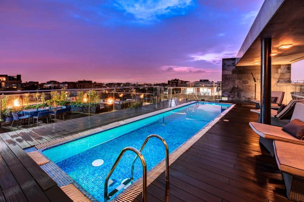 Piscina na cobertura do Hotel Estelar com espreguiçadeiras e vista para a cidade ao pôr do sol, sob um céu parcialmente nublado com tons de roxo e rosa. Imagem para ilustrar post sobre onde ficar em Bogotá.
