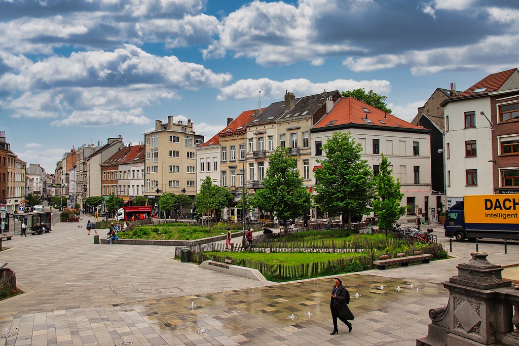 Place Fernand Cocq, em Ixelles, com pessoas caminhando, árvores, prédios modernos e um céu nublado acima.