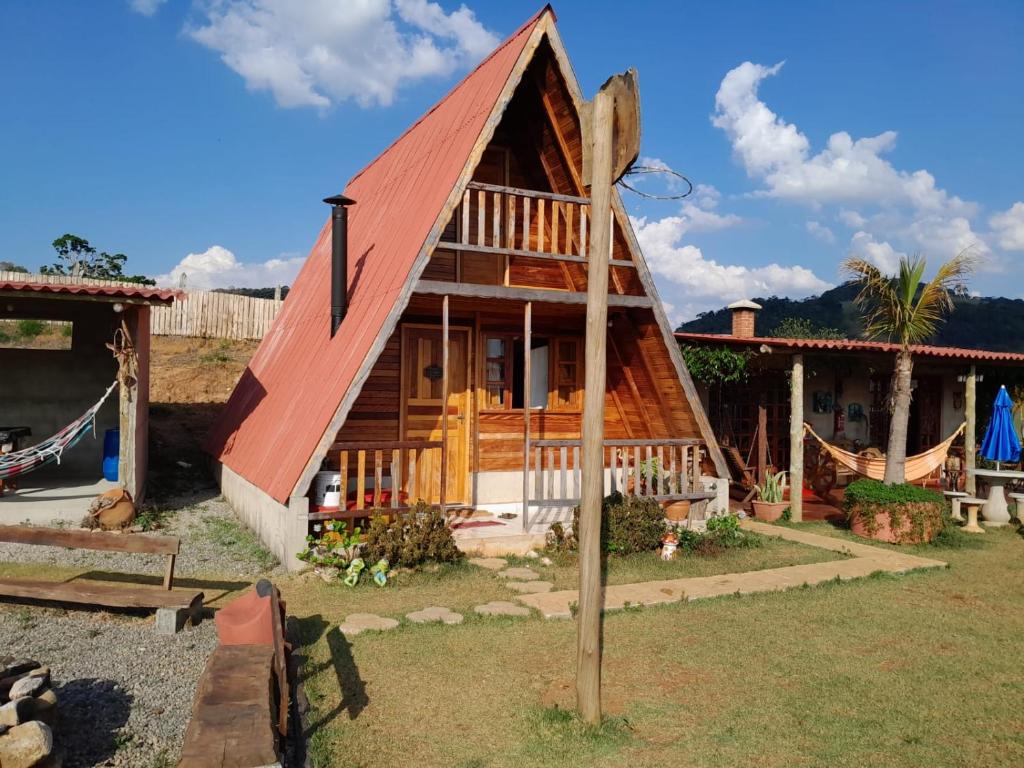 Uma cabana de madeira da Pousada e Camping Tio Will em formato de A com telhado vermelho, rede e área de jardim sob um céu azul com nuvens dispersas.