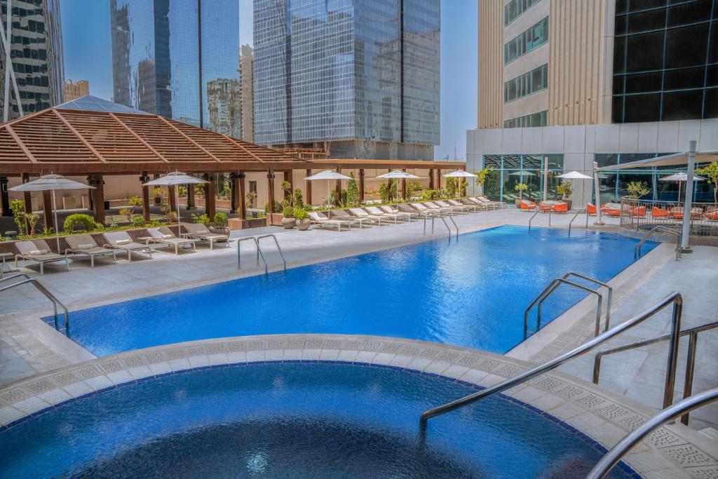 Área da piscina externa no Pullman Doha West Bay com espreguiçadeiras, guarda-sóis, um espaço coberto para sentar e prédios altos ao fundo.