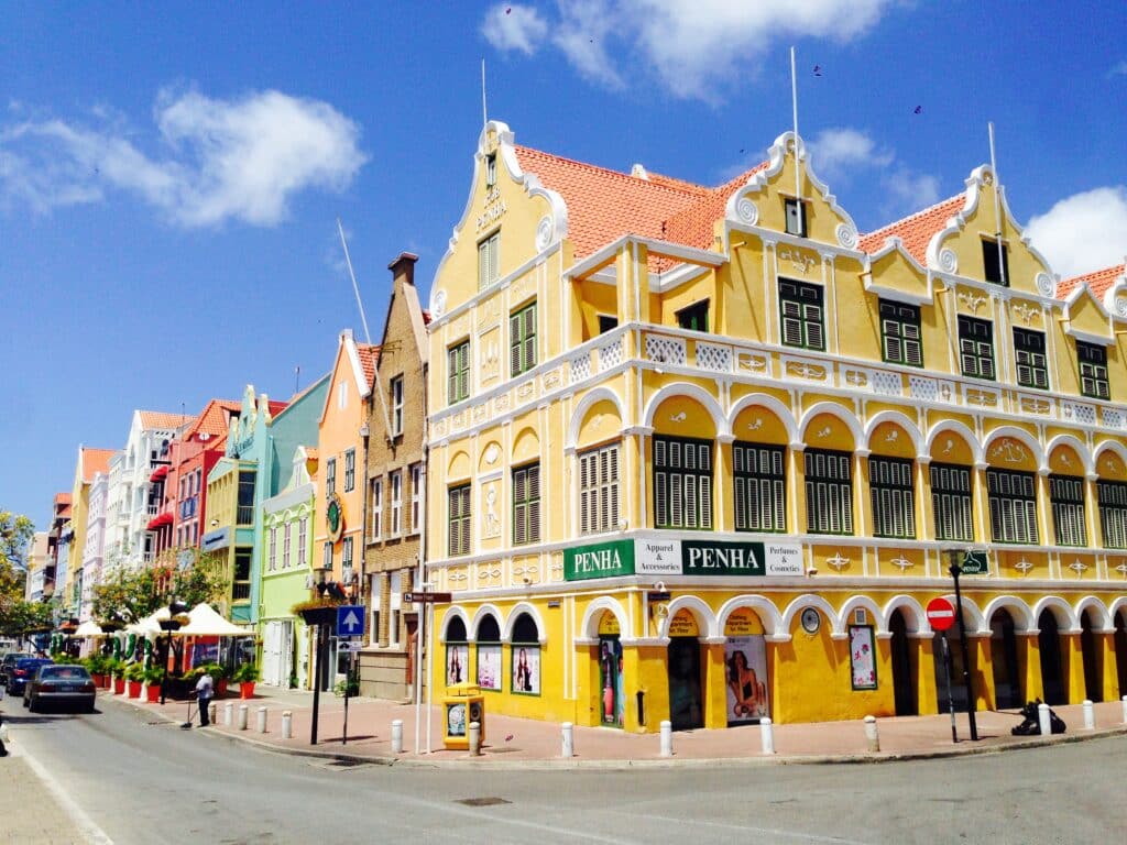 Fileira de edifícios coloniais holandeses coloridos em Punda com telhados de duas águas sob um céu azul em um dia ensolarado, com a rua e os carros visíveis. Imagem para ilustrar post sobre onde ficar em Curaçao.