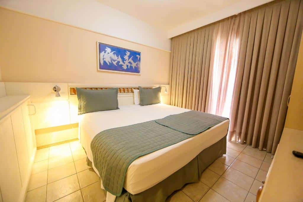 Um quarto do Destino Floripa Jurerê arrumado com uma cama de casal, roupa de cama verde, arte na parede acima da cama e cortinas bege cobrindo uma janela. Representa uma das pousadas em Jurerê Internacional.