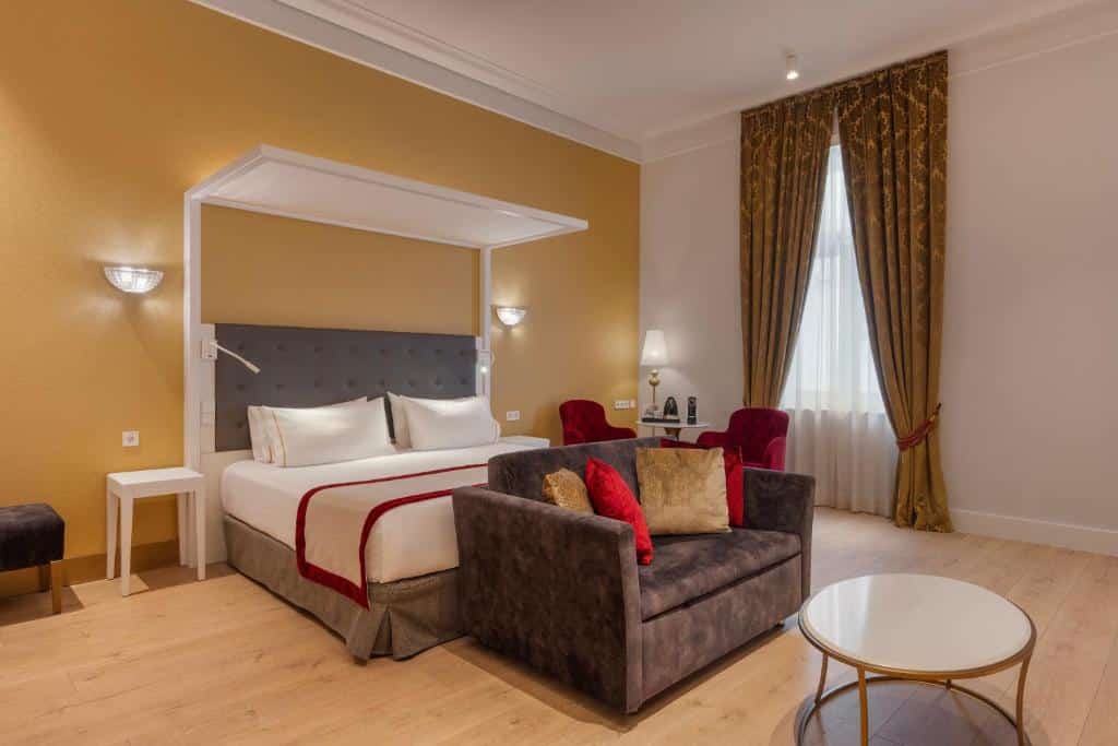Quarto do Áurea Ana Palace by Eurostars Hotel Company com uma cama de casal, sofá, mesa de centro redonda, duas cadeiras vermelhas e uma janela com cortinas douradas.