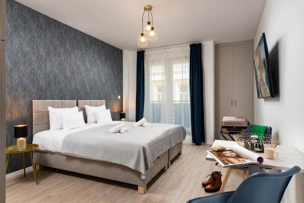 Quarto do Bo18 Hotel Superior com uma cama de casal, parede com detalhes em azul, TV, pequena escrivaninha, mala e janela com cortinas.