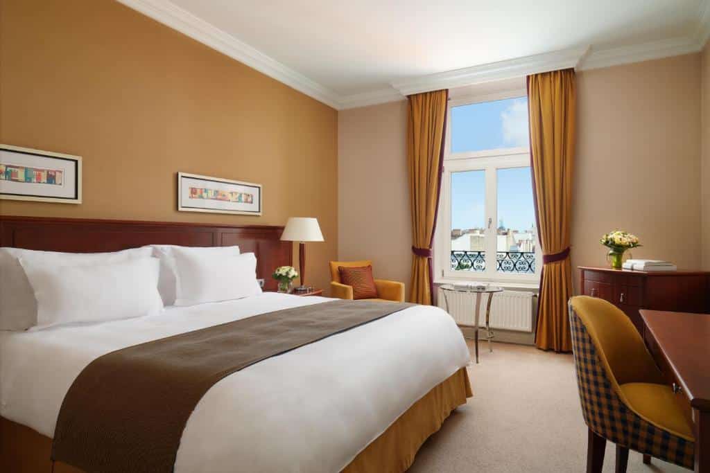 Quarto do Corinthia Budapest com uma cama grande, escrivaninha, poltrona, janela com vista para a cidade e decoração marrom-amarelada.