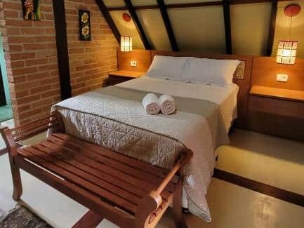 Uma cama bem arrumada do quarto da Das SteinHaus, uma das pousadas em Extrema, com duas toalhas, banco de madeira e iluminação aconchegante em um quarto aconchegante em estilo sótão com detalhes em tijolo e madeira.