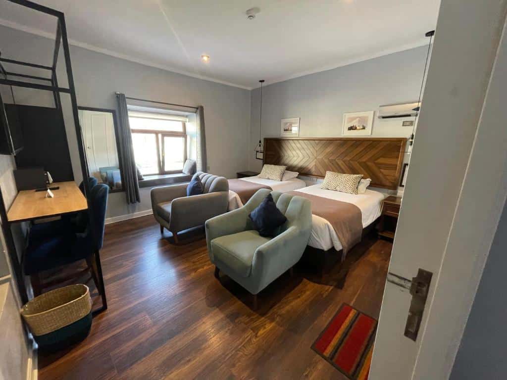 Quarto da Casa Montes San Blas hotel com duas camas, duas poltronas, uma escrivaninha, TV, janela, cesto e piso de madeira.