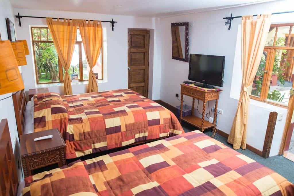 Quarto da Hosteria de Anita com duas camas de casal, colchas estampadas, TV, móveis de madeira e janelas com cortinas laranja.