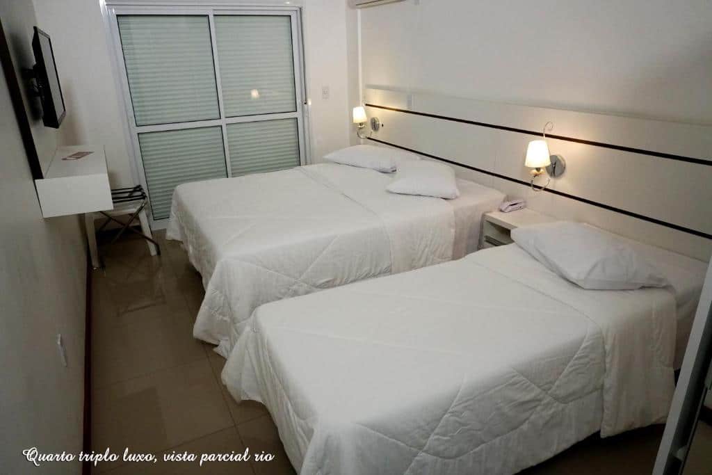 Um quarto da Pousada Molhes da Barra com duas camas de casal, roupa de cama branca, luminárias de parede, uma TV e uma janela grande com persianas fechadas.