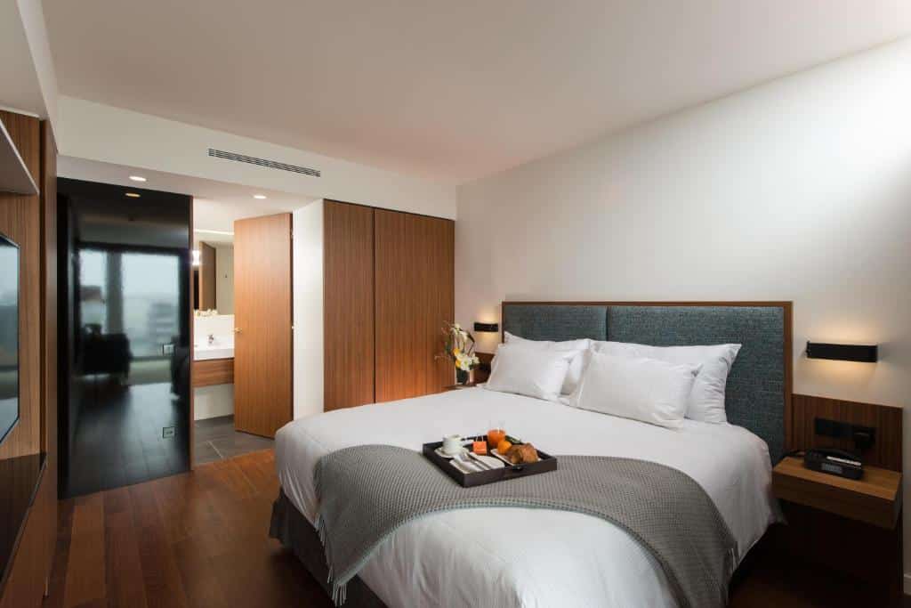 Quarto do Fraser Suites Geneva com uma cama grande, piso de madeira, uma bandeja de comida e uma porta aberta para o banheiro.