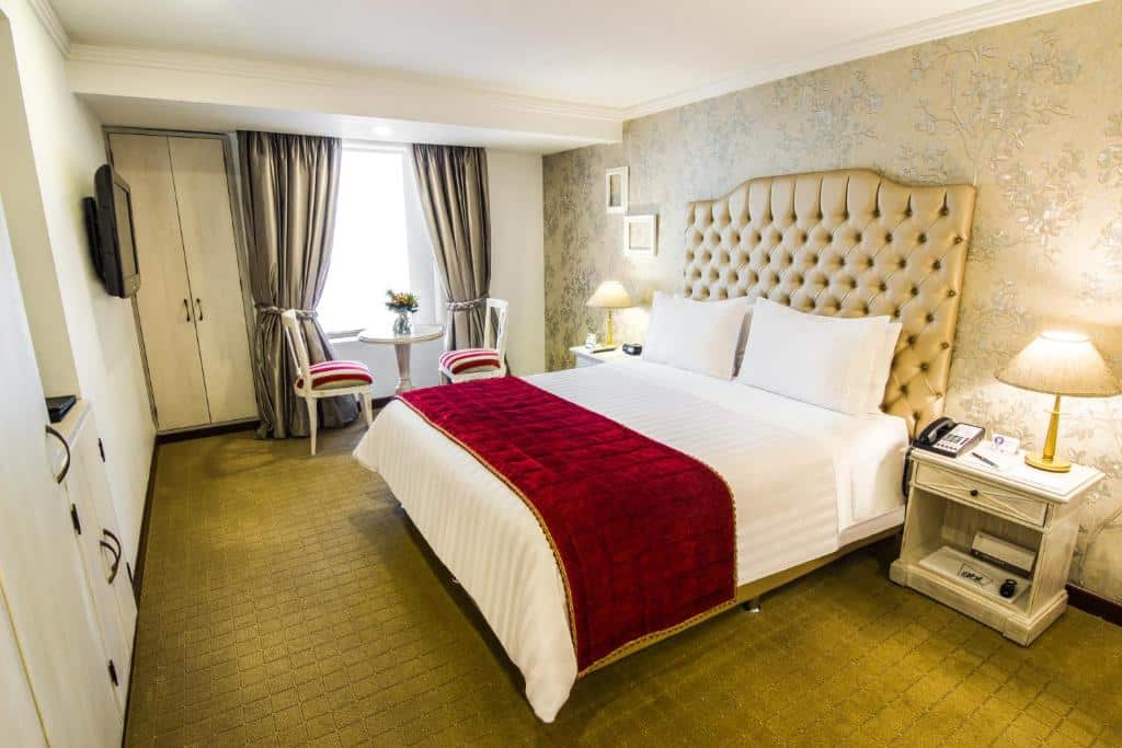 Quarto do GHL Hotel Hamilton com uma cama grande, cobertor vermelho, duas cadeiras perto de uma janela e mesas de cabeceira com lâmpadas.