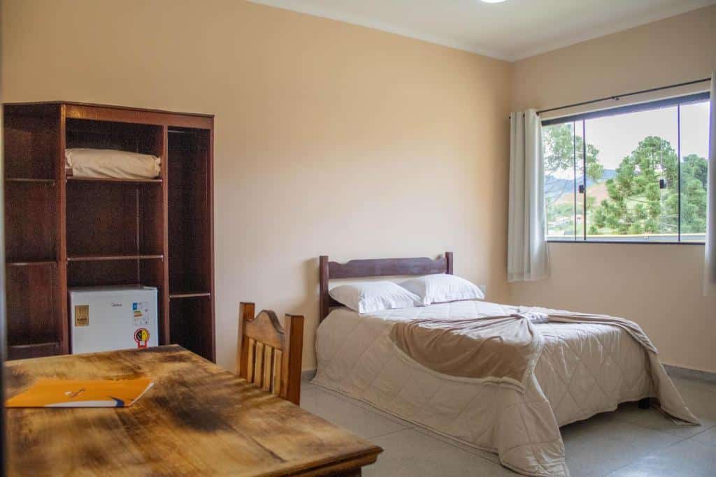 Um quarto simples do Hotel Águas Lindas com uma cama de solteiro, escrivaninha e cadeira de madeira, guarda-roupa aberto, frigobar e uma janela com vista para o exterior.