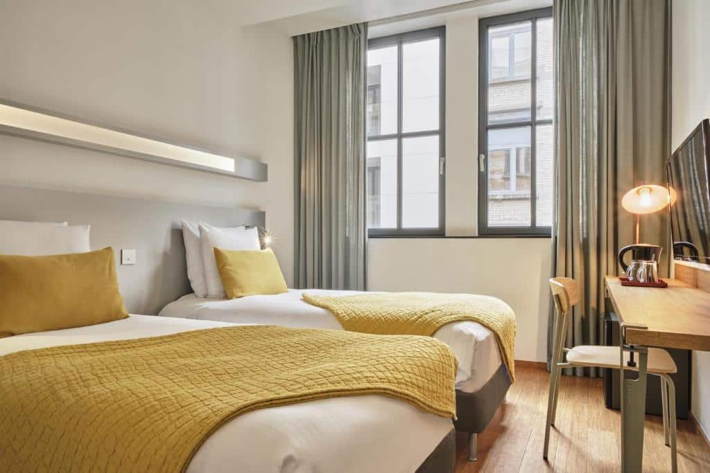 Quarto do Hôtel Aqua by HappyCulture com duas camas de solteiro, roupa de cama amarela, escrivaninha, cadeira, luminária, janela com cortinas e piso de madeira clara.