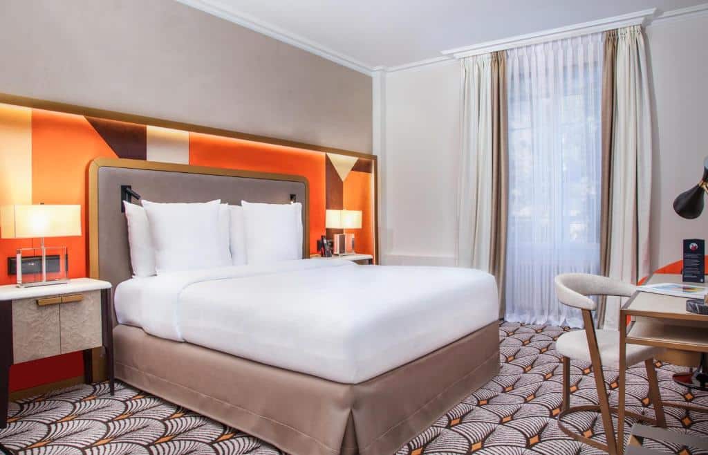 Quarto do Hotel Bristol com uma cama grande, duas mesas de cabeceira com luminárias, uma escrivaninha com uma cadeira e uma janela com cortinas transparentes.