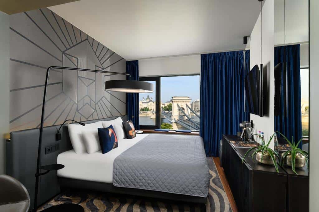 Quarto do Hotel Clark com uma cama grande, cortinas azuis, janela com vista para a cidade, arte na parede e uma cômoda com plantas e amenidades.