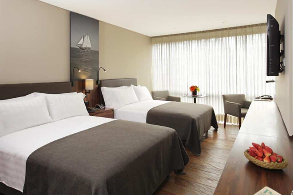 Quarto do Hotel Estelar com duas camas de casal, uma TV na parede, uma mesa com duas cadeiras e uma janela grande com cortinas transparentes.