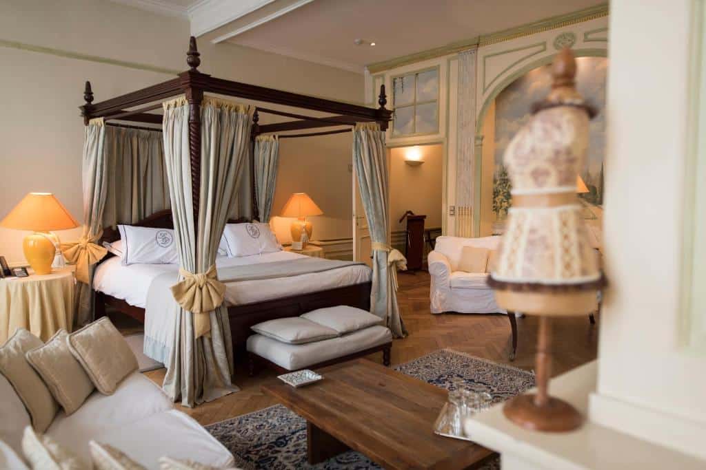 Quarto espaçoso do Hotel Le Dixseptième com uma cama de dossel, sofá, poltronas, mesa de madeira, luminárias e uma forma de vestido em primeiro plano.