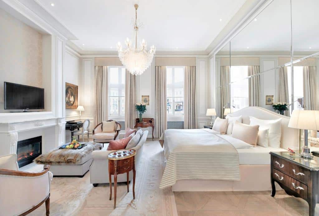 Quarto do Hotel Sacher Wien elegante com cama, sofá, lustre, lareira, TV, janelas grandes e parede espelhada, para representar onde ficar em Viena.