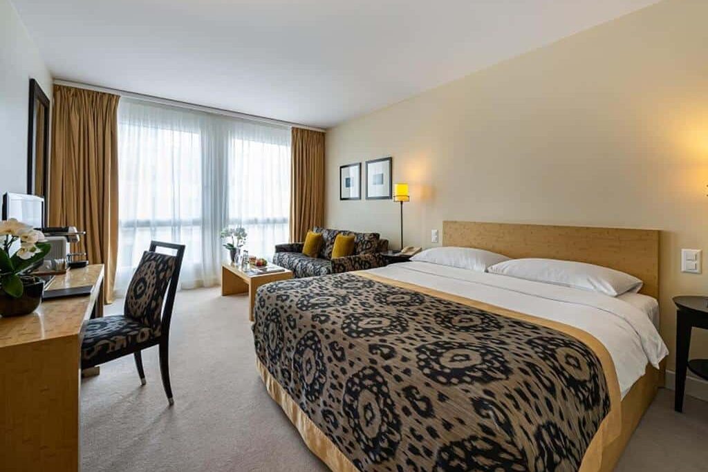 Um quarto do Jade Manotel com uma cama de casal, escrivaninha, cadeira, sofá, janela grande com cortinas e decoração em cores neutras.