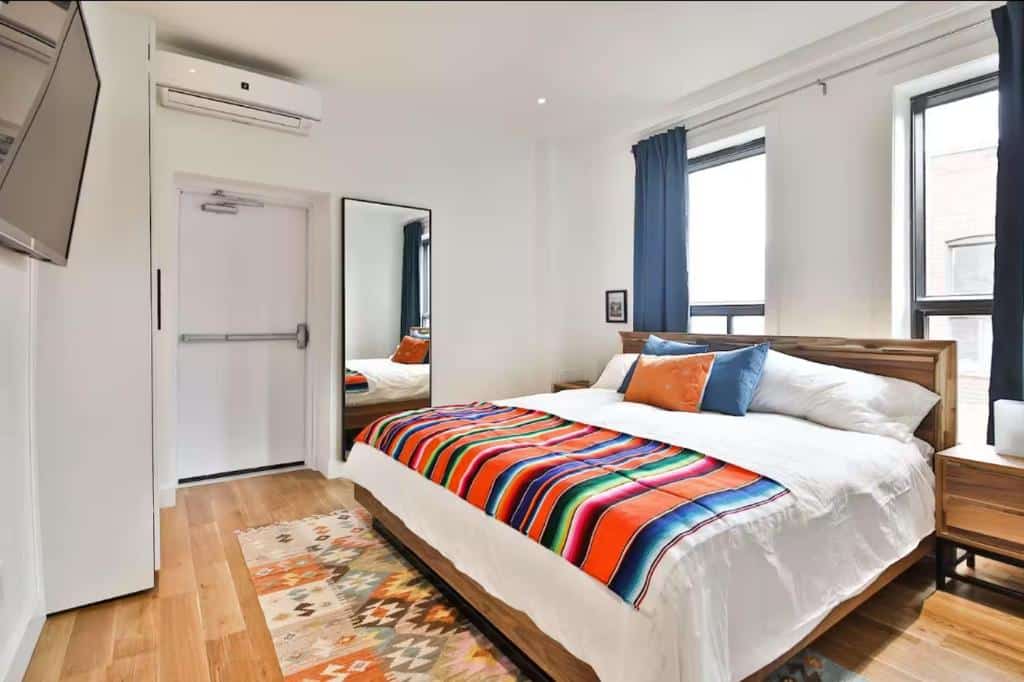 Quarto do Les Lofts du Mile End-Plateau Mont Royal bem iluminado com uma cama king size, cobertor listrado colorido, tapete estampado, espelho grande e duas janelas com cortinas azul-marinho.