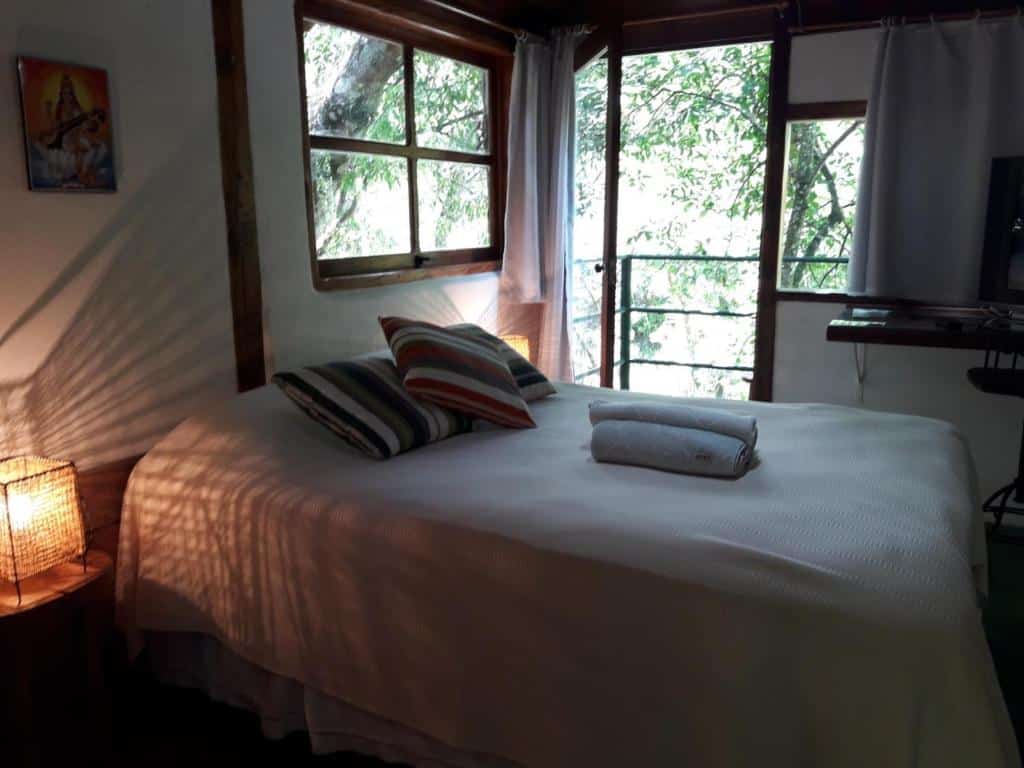 Uma cama com travesseiros listrados e toalhas fica ao lado de janelas e uma porta de vidro, com árvores visíveis do lado de fora e iluminação interna suave, da Pousada Estância Rio Acima.