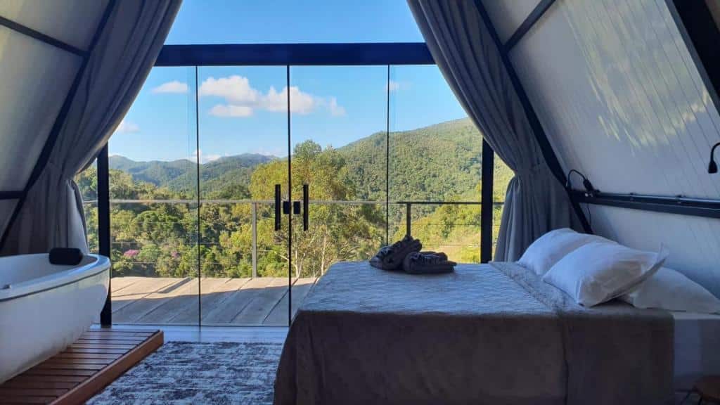 Uma cama e uma banheira em uma cabine de estrutura em A com portas de vidro que se abrem para uma varanda e uma vista das colinas verdes, da Pousada Sítio Nova União.
