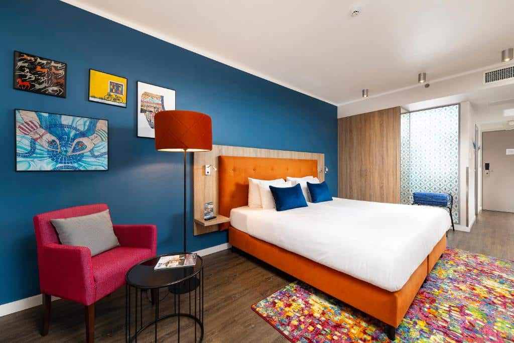 Um quarto do Three Corners Lifestyle Hotel com uma cama laranja, parede de destaque azul, cadeira vermelha, obras de arte e tapete colorido no piso de madeira.