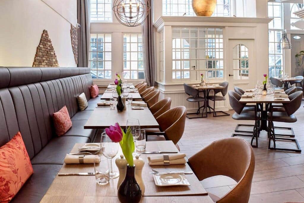 Foto do Radisson Blu Hotel. Interior de restaurante moderno com mesas de madeira, cadeiras almofadadas, vasos de tulipas, janelas grandes e tons neutros de decoração.