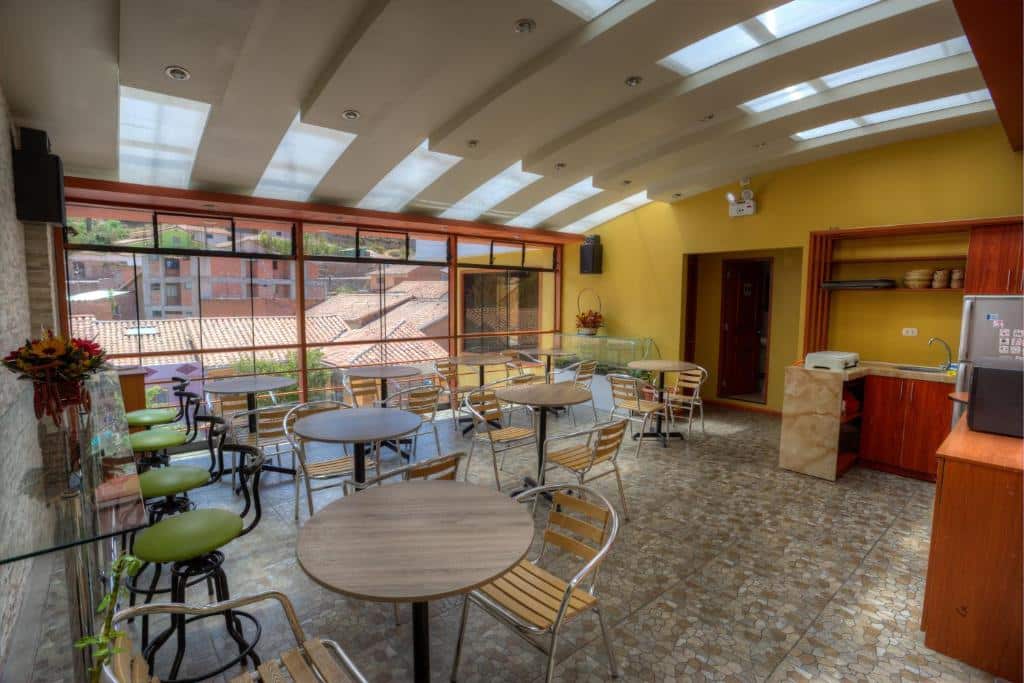 Uma sala bem iluminada do El Mariscal Cusco com mesas redondas, cadeiras, uma parede amarela, janelas grandes e um teto com claraboia.