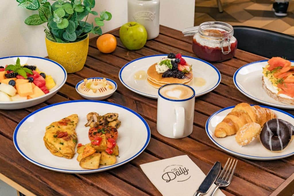 Uma mesa de madeira com pratos de panquecas, frutas, omelete, bolos, café e um pote de geleia, com uma planta e frutas. O café da manhã é do Rove Dubai Marina.